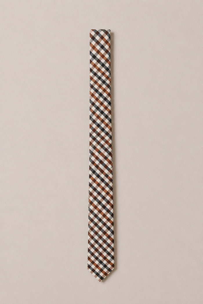 A slim cotton tie from Ivy City Co, featuring their Ties in Chestnut Gingham design—showcasing a classic plaid pattern in brown, black, and white—displayed against a light beige background, making it an ideal choice for a stylish wedding accessory.