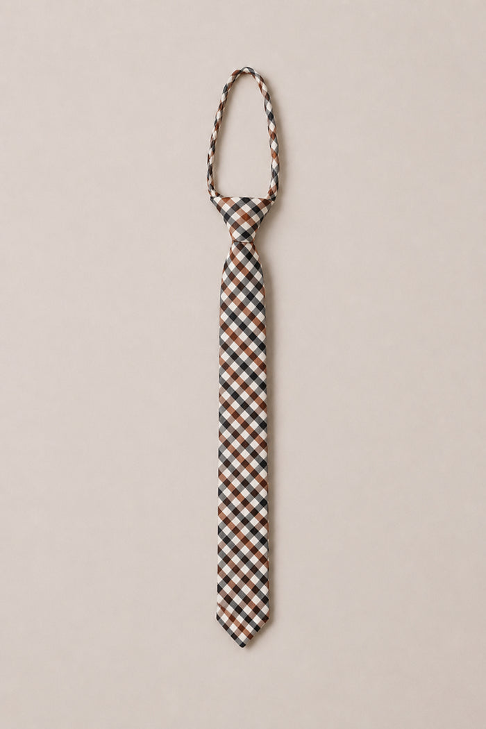 The Ivy City Co Ties in Chestnut Gingham, featuring a brown, black, and white plaid pattern, is showcased on a simple light beige background—a perfect cotton tie to elevate your wedding attire with style.