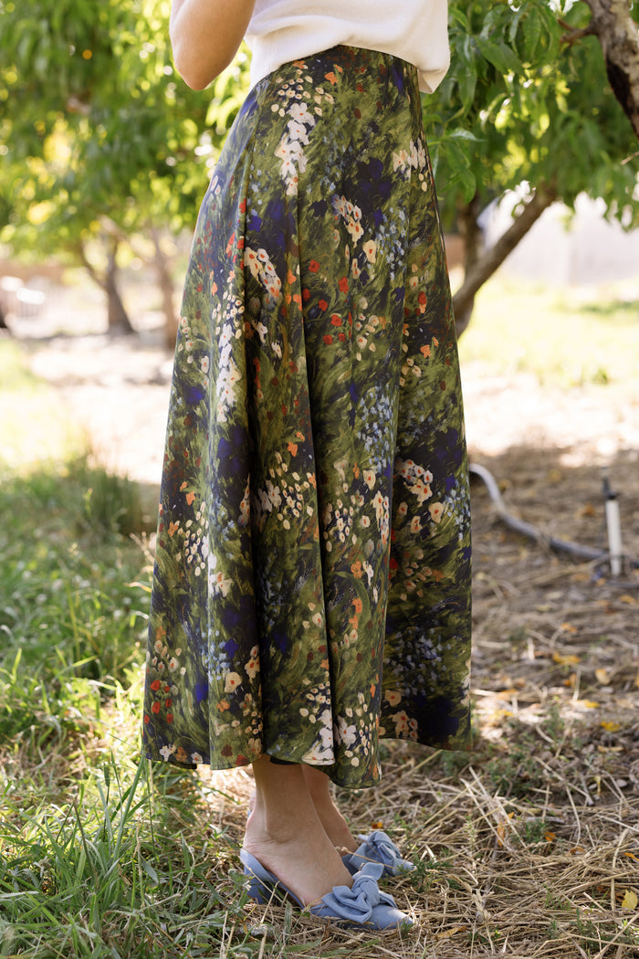 A woman stands outdoors surrounded by grass and trees, wearing a white top paired with the Ivy City Co Tiff Skirt in Impressionist Bloom, and blue bow flats. Her face is not visible in the image.