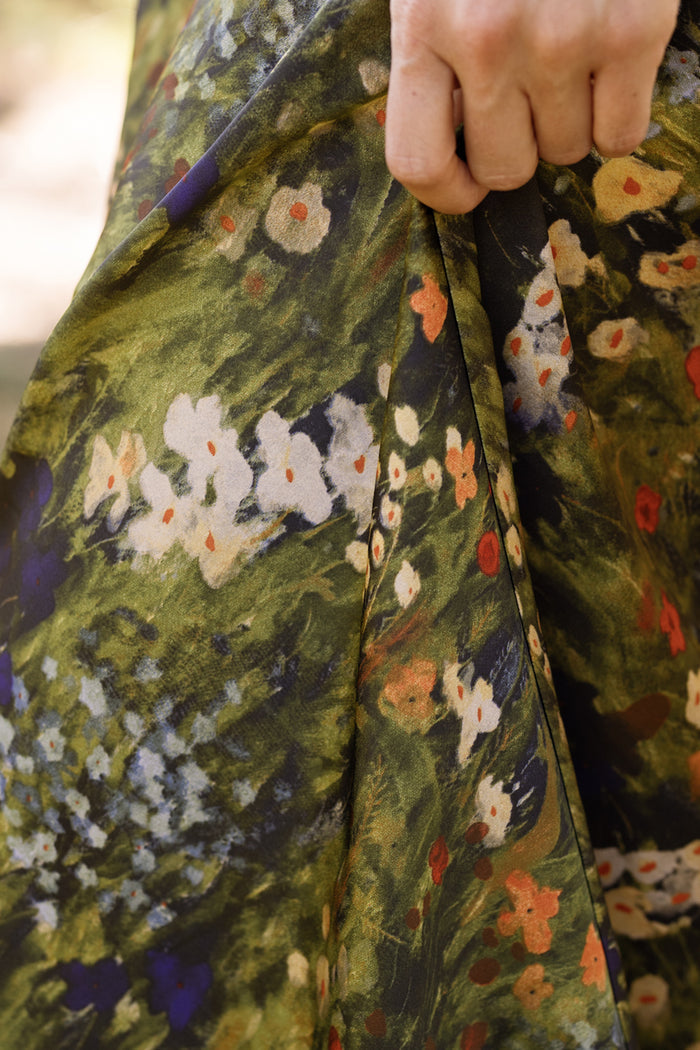 A close-up of a hand holding a green fabric with a colorful floral pattern featuring white, orange, and blue flowers—the same vibrant satin used for Ivy City Co’s Tiff Skirt in Impressionist Bloom, perfect for your next night out.