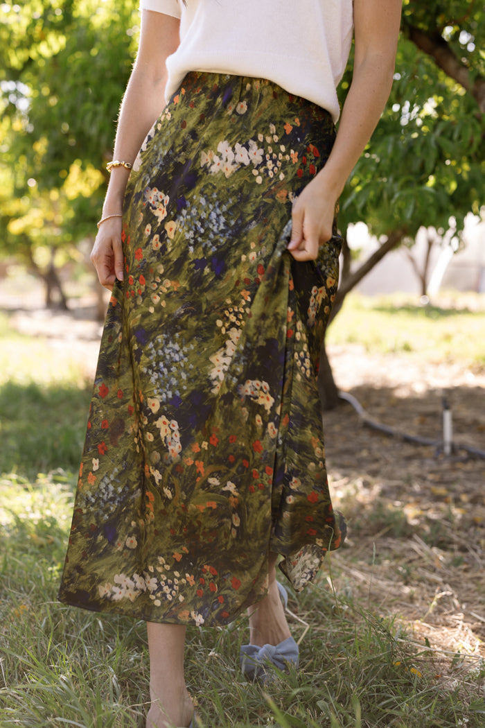 Outdoors, a person stands on grass beneath trees and sunlight, gently holding the Tiff Skirt in Impressionist Bloom by Ivy City Co, styled with a white top.