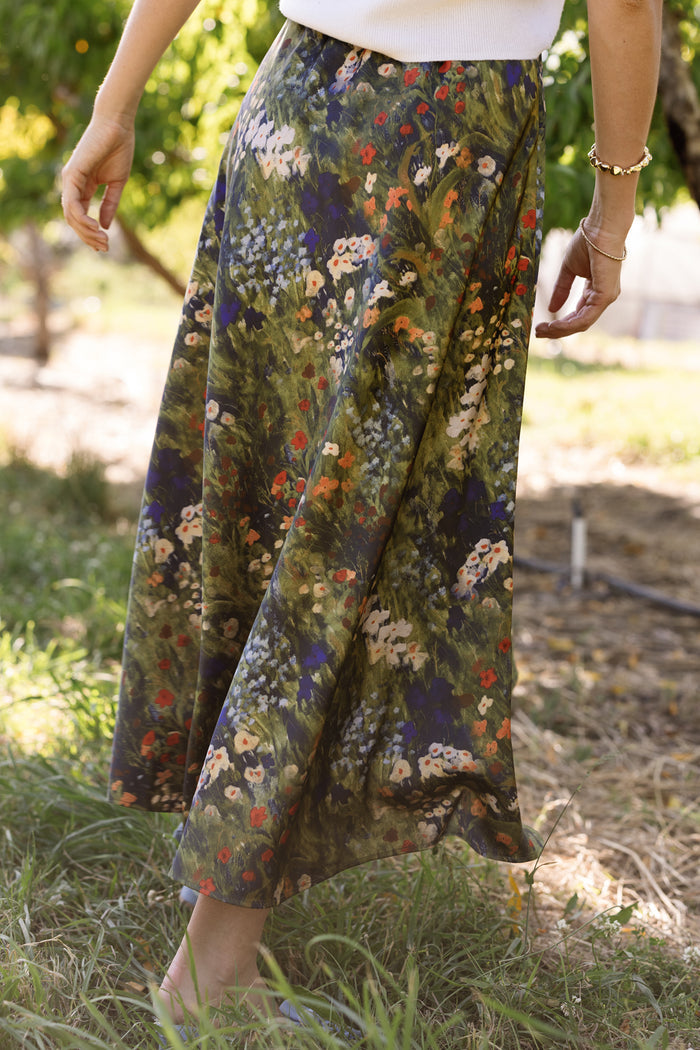 A person wearing the Ivy City Co Tiff Skirt in Impressionist Bloom, a long and flowy satin skirt featuring a colorful floral print, stands barefoot on grass outdoors in a sunlit garden or park.