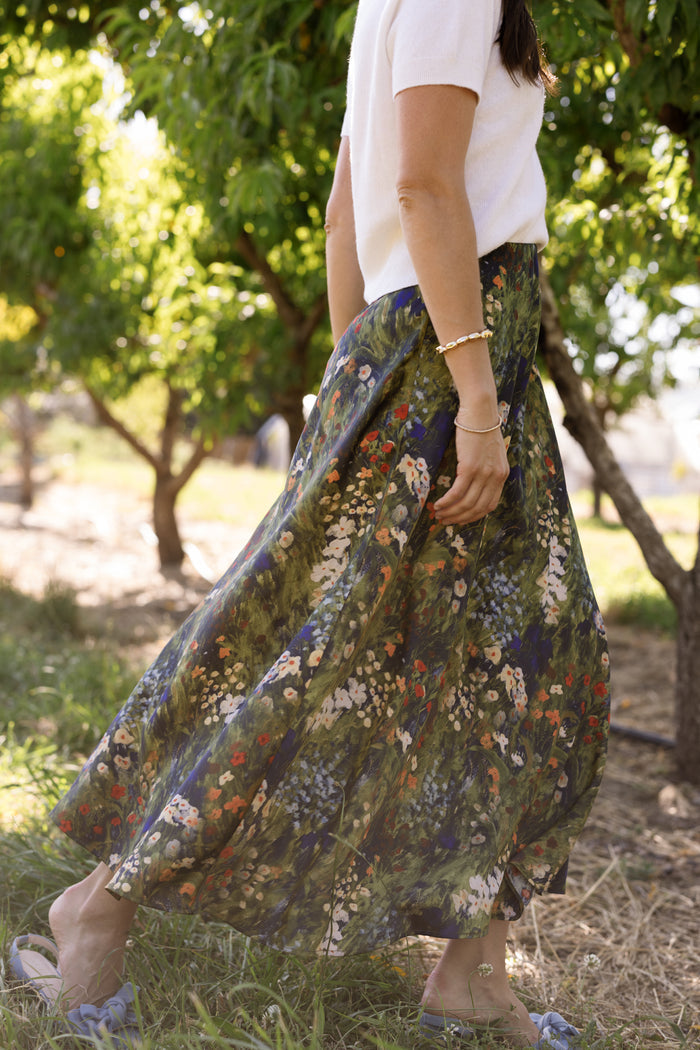 A woman stands outdoors among trees, sunlight filtering through the green leaves as she wears a white short-sleeve top paired with the Ivy City Co Tiff Skirt in Impressionist Bloom, its long, flowing silhouette adorned with a delicate floral print. Only her lower face and body are visible.