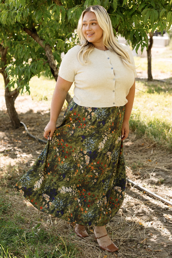 A woman with long blonde hair smiles while standing outdoors near leafy trees, wearing a short-sleeve beige top paired with Ivy City Co’s Tiff Skirt in Impressionist Bloom, which features a flowy, floral pattern.