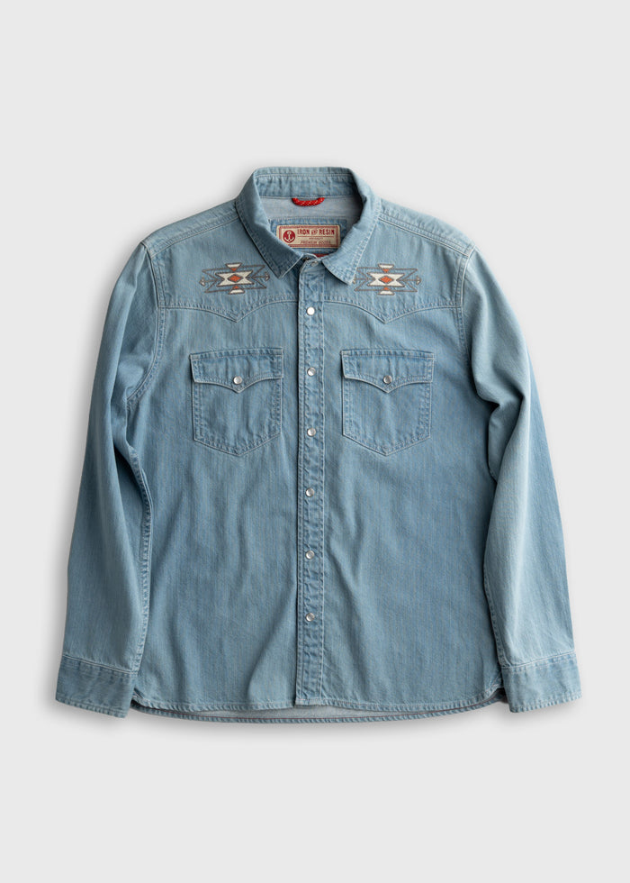 Light blue denim shirt with embroidered geometric patterns on shoulders, buttoned front, and two chest pockets on a plain background.