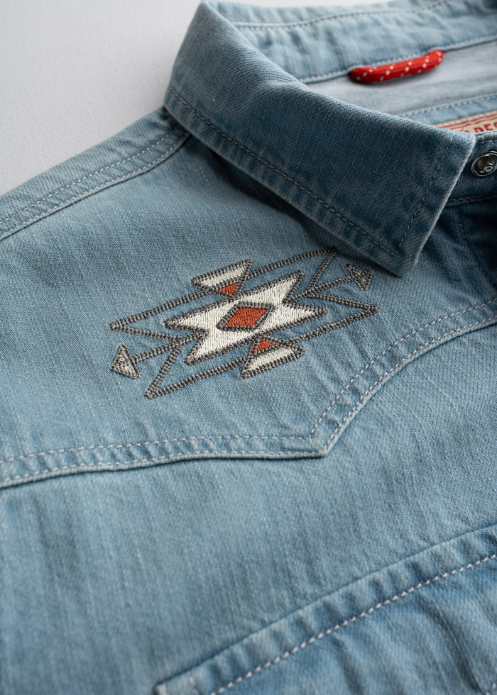 Close-up of a denim shirt with a geometric embroidered pattern near the collar, featuring red and white accents.