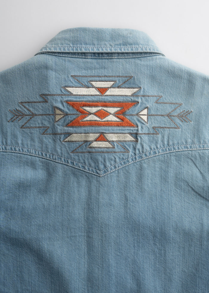 Denim shirt back with geometric embroidery in orange, white, and gray, featuring arrows and triangles, on a light background.