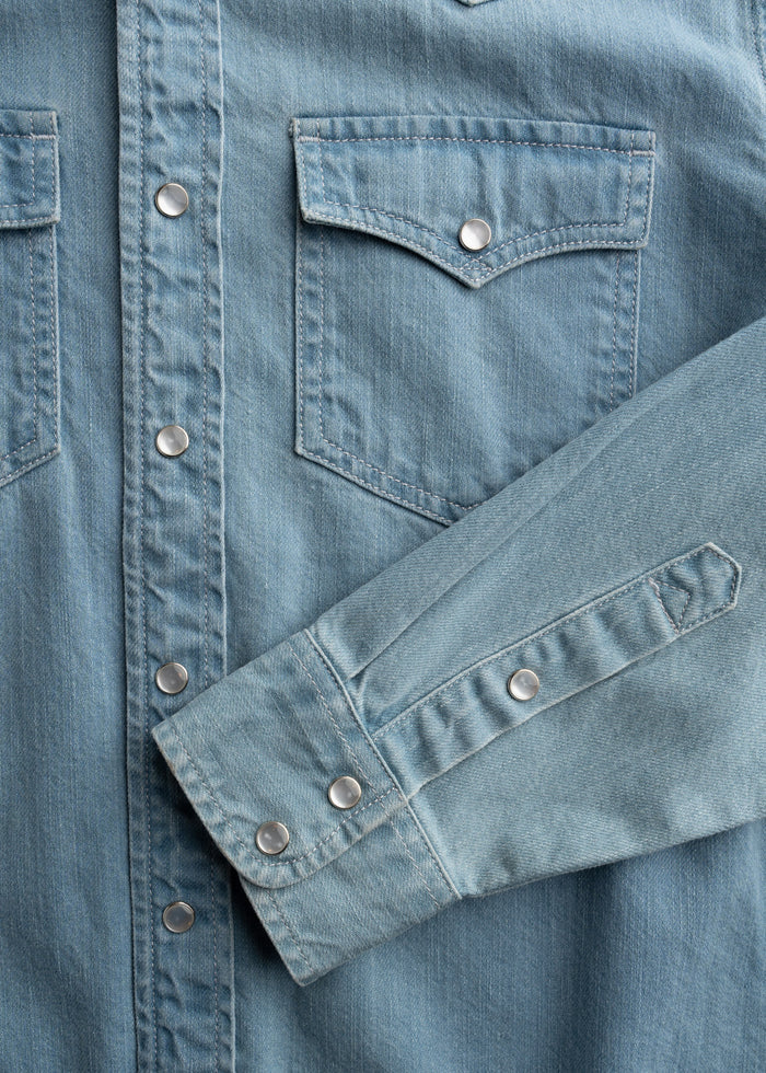 Close-up of a blue denim shirt with metal snap buttons and two chest pockets, highlighting the fabric texture and stitching.