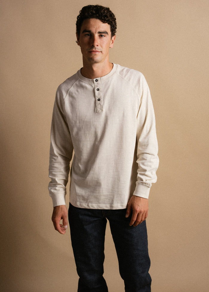 Man in a cream henley shirt and dark jeans stands against a beige background, looking confidently at the camera.