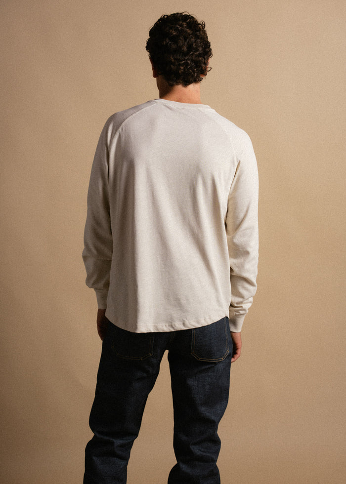 Man in a white long-sleeve shirt and jeans stands with back to camera against a beige background, highlighting casual fashion.