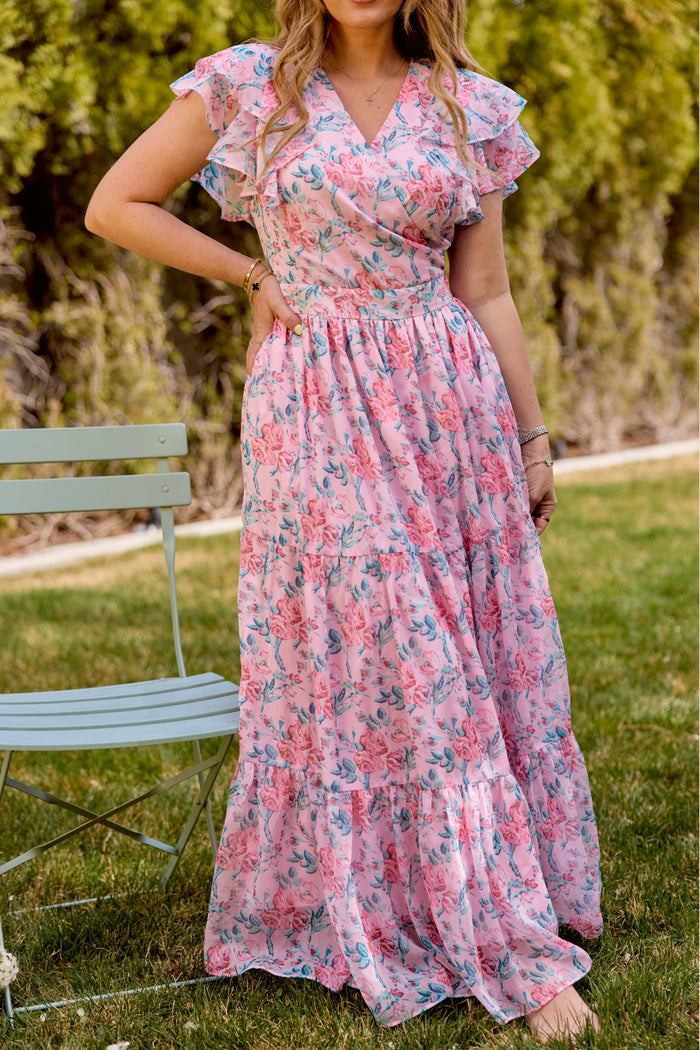 A woman in the Trixie Dress in Pink Sketched Flower Chiffon by Ivy City Co stands on grass near a pale green chair, framed by leafy trees. Her face is hidden—ideal inspiration for a summer wedding look.