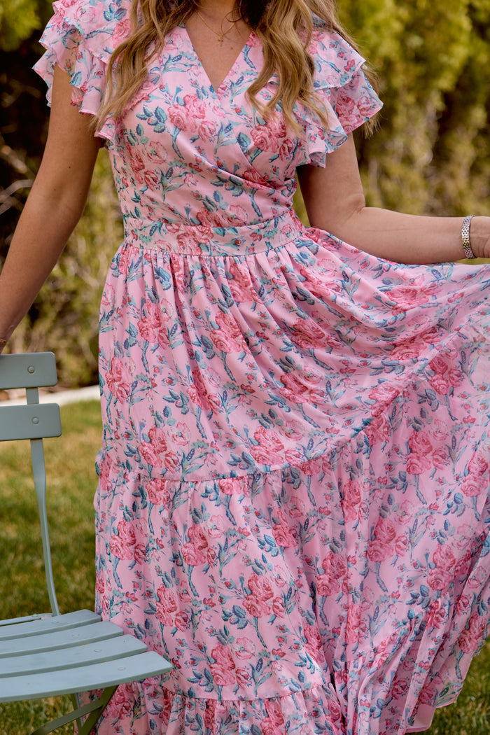A woman in the Ivy City Co Trixie Dress in Pink Sketched Flower Chiffon stands outdoors, holding out the airy midi skirt by a light blue chair and lush greenery—her pose highlights this perfect summer wedding look.