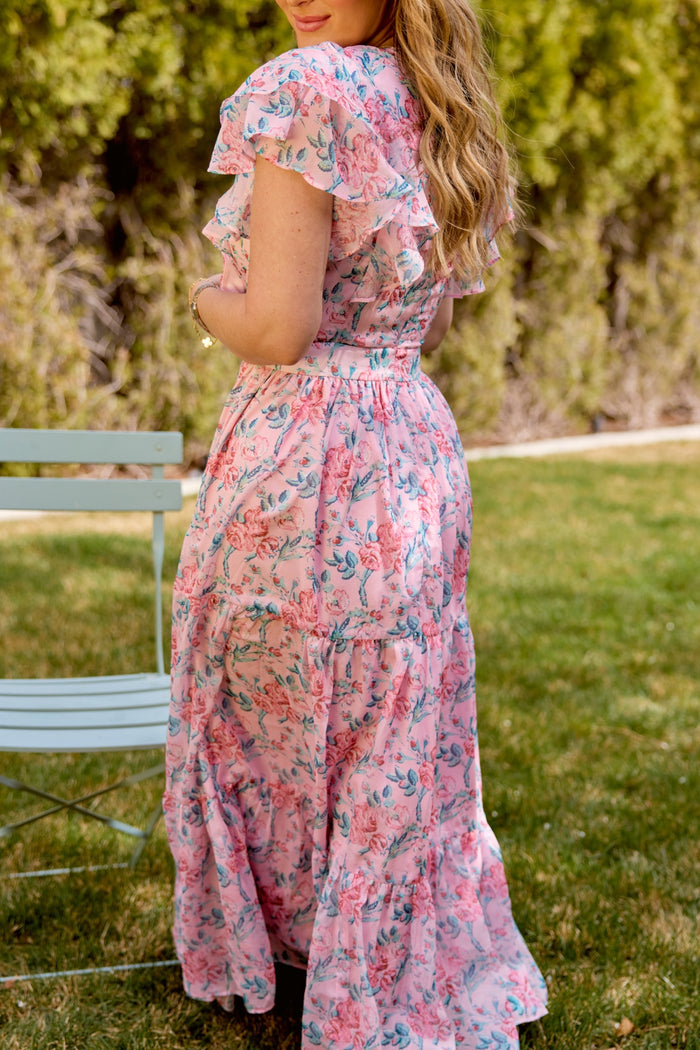 A woman with long wavy hair wears the Ivy City Co Trixie Dress in Pink Sketched Flower Chiffon, featuring ruffled sleeves and a tiered skirt, standing outdoors by a light blue chair—a perfect look for a summer wedding.