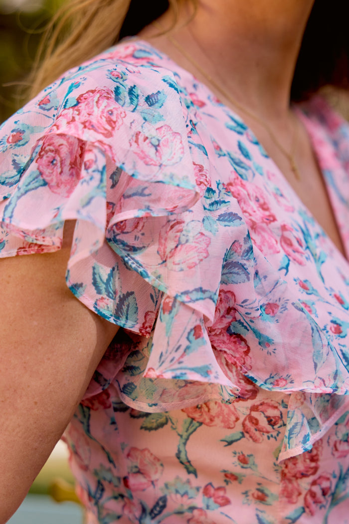 Close-up of a person wearing the Ivy City Co Trixie Dress in Pink Sketched Flower Chiffon, featuring ruffled short sleeves and pink and blue floral print—perfect for summer weddings. A gold necklace adorns their neck.