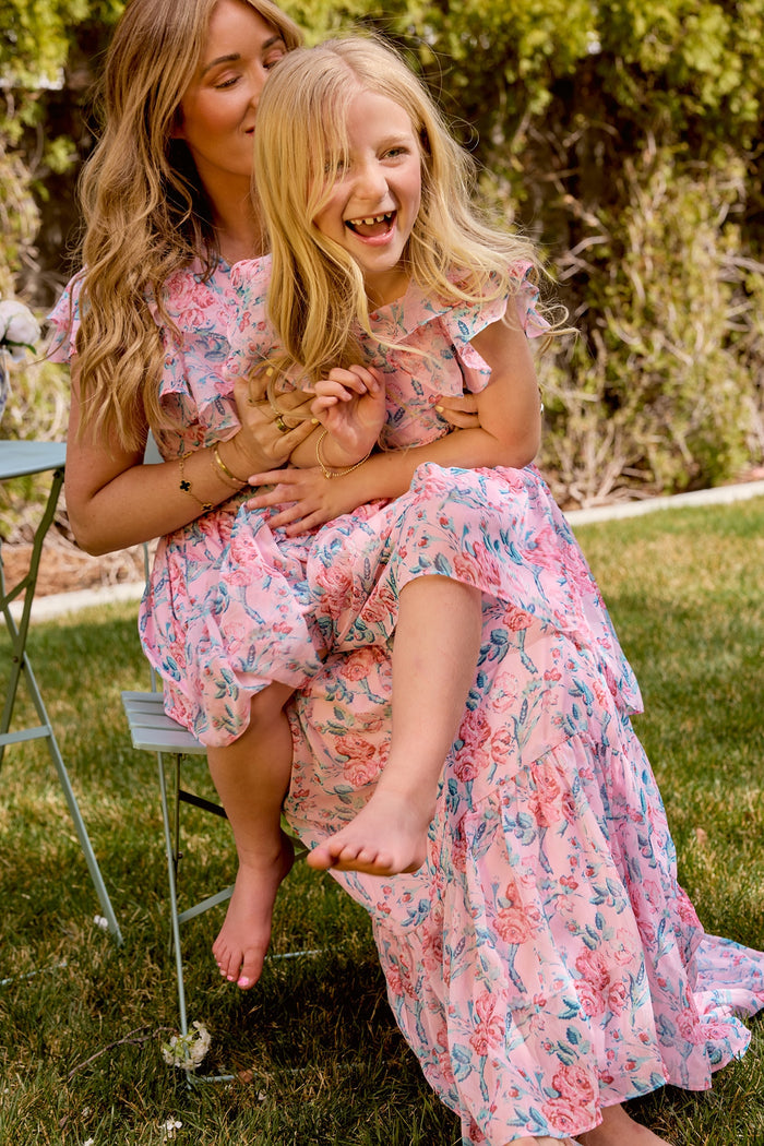 A woman and young girl sit together in a garden, both smiling and relaxed in matching Ivy City Co Trixie Dresses in Pink Sketched Flower Chiffon—perfect stylish outfits for a summer wedding.