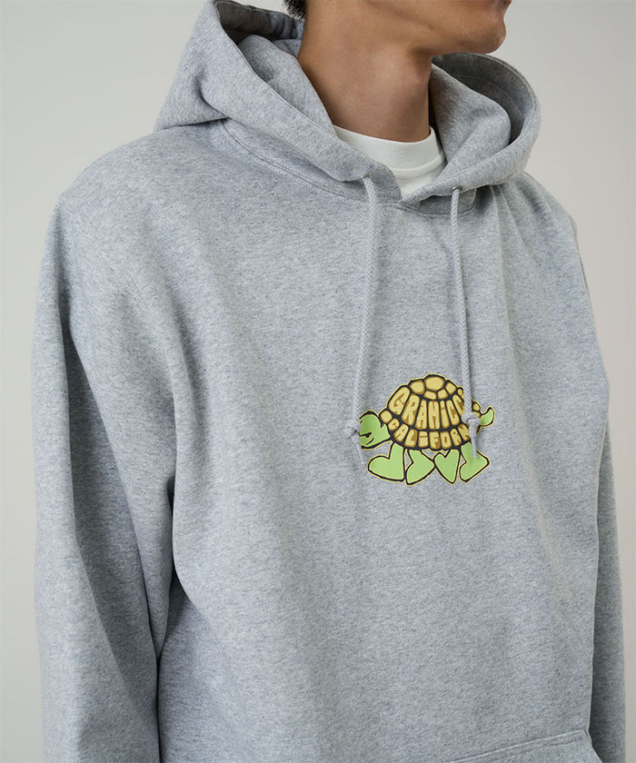 Model wearing a Gramicci heather grey Turtle Hooded Sweatshirt with turtle graphic against a light tone background.