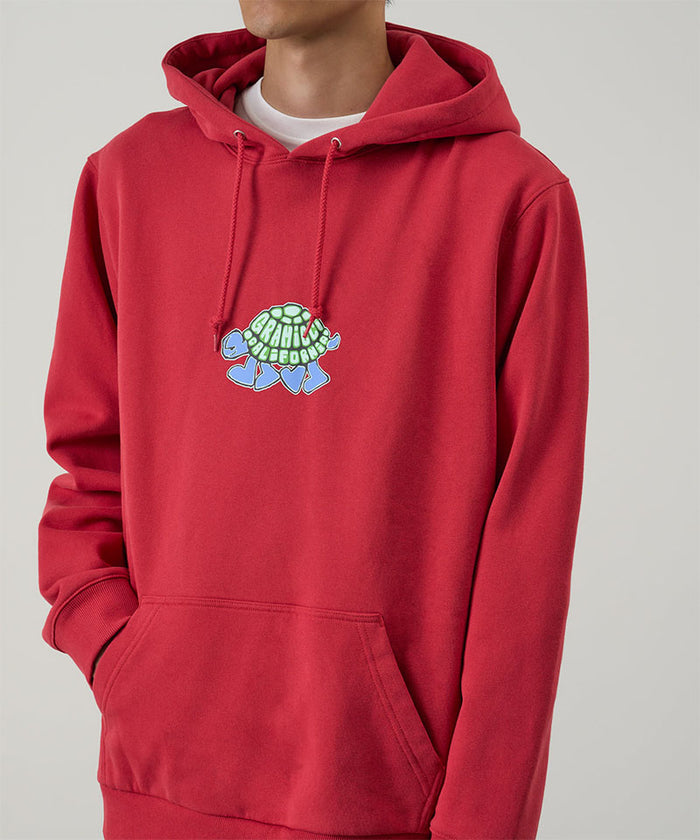Model wearing a Gramicci crimson red Turtle Hooded Sweatshirt with turtle graphic against a light tone background.