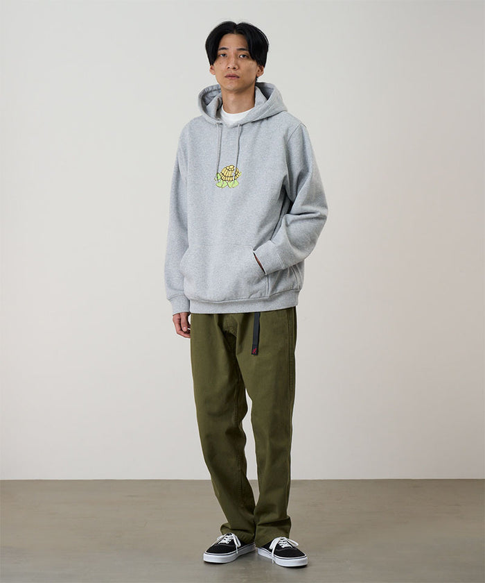 Model wearing a heather grey Gramicci Turtle Hooded Sweatshirt with a turtle graphic and olive pants on a light tone background.