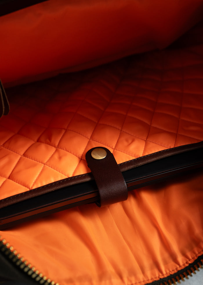 Open bag with orange quilted lining, leather strap, and brass button, showcasing a sleek, secure compartment for a laptop.