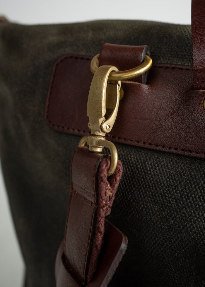 Close-up of a brown leather bag strap with a brass clasp attached to a dark canvas bag, highlighting craftsmanship details.