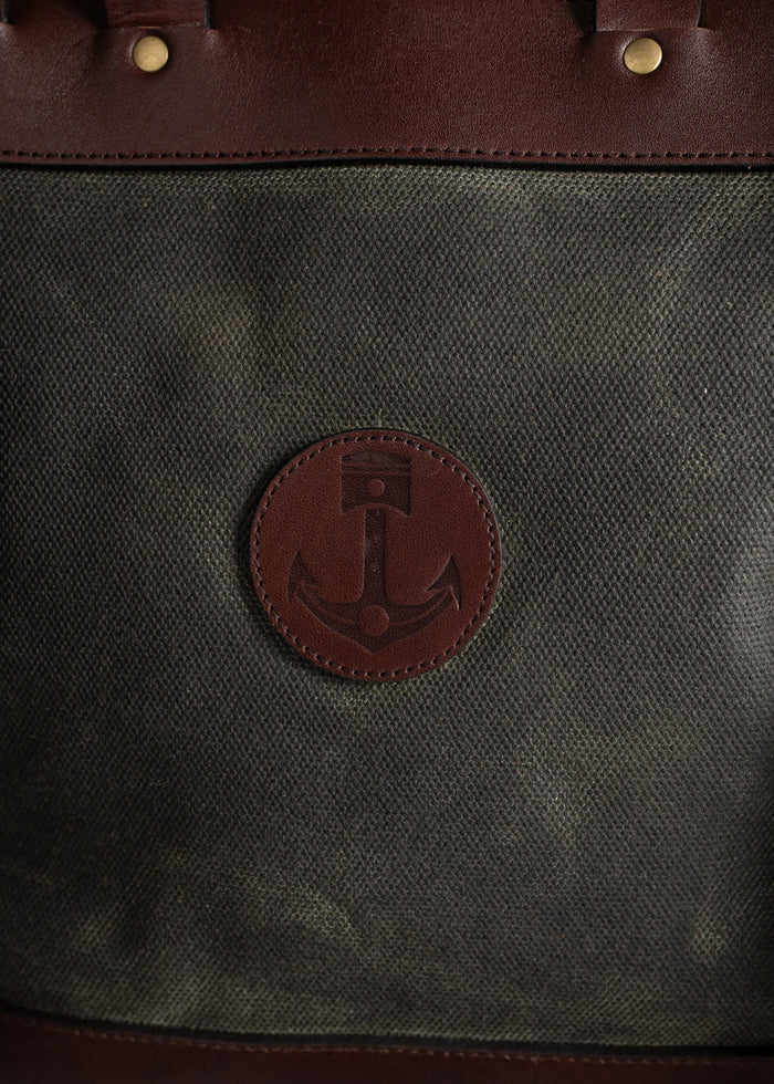 Brown leather bag with anchor emblem on canvas fabric, featuring brass rivets. Classic nautical design and sturdy craftsmanship.