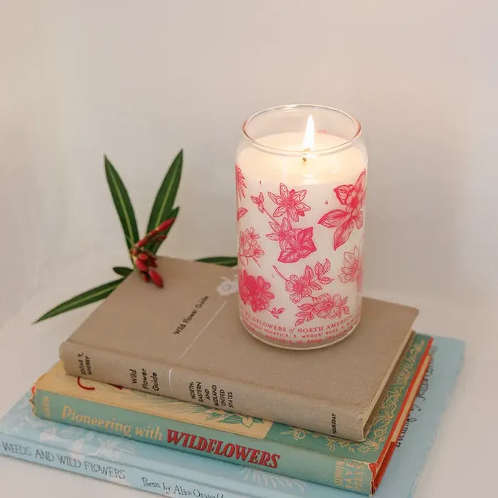 Lit Woodland Wildflowers Soy Candle in a pink floral glass jar atop a stack of vintage books with a small plant nearby.