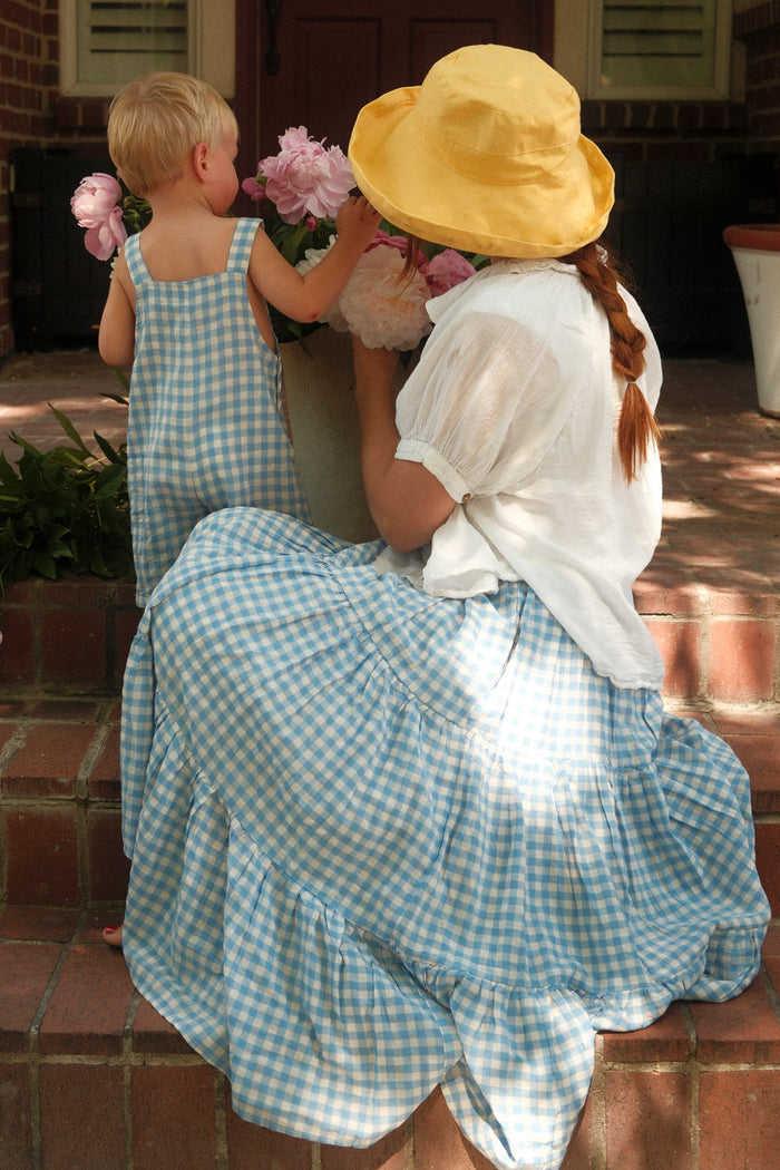 A woman wearing the Ivy City Co Zoey Skirt in Blue Gingham sits on steps with a child in matching overalls. They admire pink flowers together, sharing a sweet moment in their coordinated outfits.