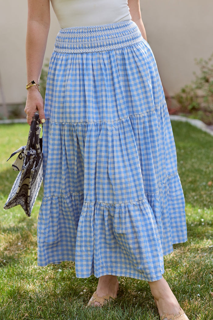 A person stands on grass wearing the Ivy City Co Zoey Skirt in Blue Gingham, styled as a maxi, and holding two patterned scarves. The upper body is mostly out of frame.