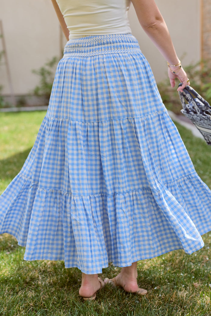 A person stands on grass, wearing the Zoey Skirt in Blue Gingham by Ivy City Co and holding a patterned bag. Only the lower body is visible.